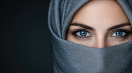 Naklejka premium Beautiful Muslim woman with striking blue eyes looks directly forward while wearing a grey hijab in a studio setting.