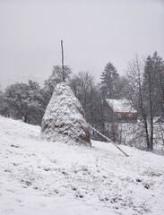 haystack on a slope, house behind trees, snowy day in a mountain village