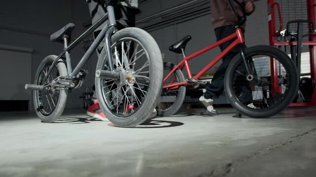Low section shot of two anonymous guys in sporty baggy clothes walking past camera in slow motion with red and black BMX bikes while training in hangar