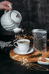 woman pouring green tea from a white teapot into a white cup with a saucer, tea drinking, dark background, wooden plate, a drop of tea falls into the cup