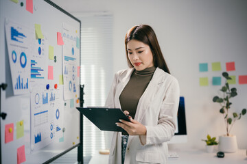 Professional analyzing project data, reviewing charts on clipboard with whiteboard metrics during...