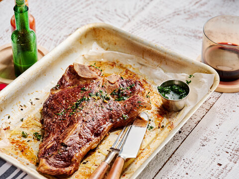 A beautifully cooked steak rests on a tray with a side of green sauce, accompanied by drinks in glasses, creating a warm and inviting dining atmosphere