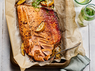 Tender baked salmon fillet topped with herbs and lemon slices, served on parchment paper alongside refreshing drinks on a rustic wooden table
