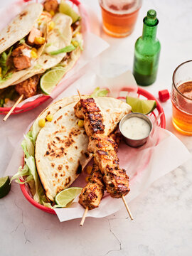 A vibrant outdoor setting features grilled chicken skewers with fresh tacos filled with vegetables. Ice-cold drinks accompany the delicious meal, creating a festive atmosphere