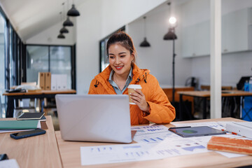Young asian woman wearing orange shirt hold use laptop pc computer drink coffee sit alone at table...