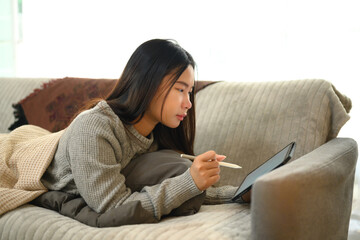 Young woman lying comfortably on the couch and using digital tablet with a stylus pen