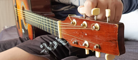 A teenage boy is sitting on the couch and tuning guitar strings. Guitar lesson. Musical instrument....
