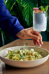 A woman's hand elegantly holds a refreshing cocktail, while a vibrant and colorful plate of fresh salad sits on a wooden table