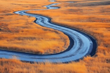 Fototapeta premium Winding wet asphalt road resembling a snake meanders through a dry grass field, bathed in the warm glow of a sunset
