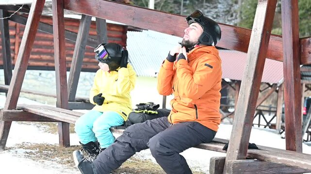 A man and a boy, bundled in warm ski gear, sit on a wooden bench outdoors. They adjust their helmets, preparing for a day on the slopes. The snowy landscape is visible in the background.