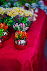 Vaso decorativo metálico redondo com arranjo de flores artificiais coloridas em tons de laranja e verde sobre toalha vermelha com textura floral em ambiente de exposição