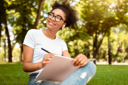 Smiling Black Studet Girl Sketching Learning To Draw Sitting In Green Park Outside. African American Millennial Female Taking Notes Looking Aside Posing Outdoors. Studentship Concept