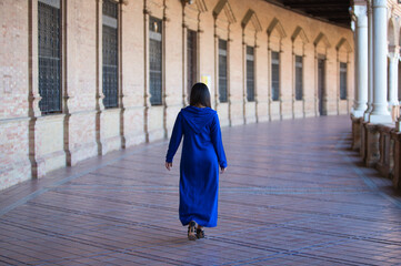 Naklejka premium moroccan brunette woman in blue abaya, typical arabian costume, walking between arches and columns in the spain square in seville. Photo taken from behind. Travel and holiday concept.