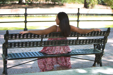 a young, beautiful brunette woman is sitting on a park bench, relaxed with her arms resting on the...