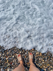 Female feet in sandals on pebble beach with sea foam