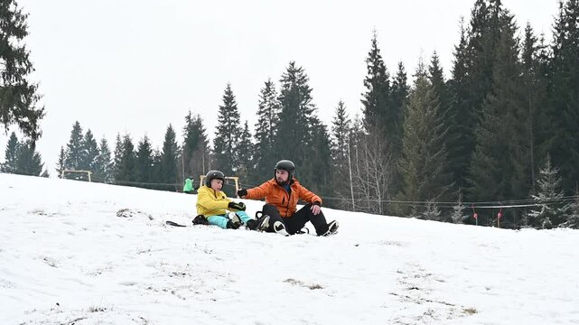 A man and child in winter gear pause on a snowy slope, after a snowboarding run. Pine trees form a backdrop, suggesting a mountain setting.