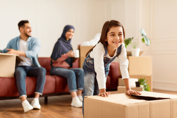 House Move And Relocation Concept. Portrait Of Cheerful Muslim Family Housing, Selective Focus On Happy Smiling Little Girl Moving Cardboard Box On Floor, Parents Sitting With Open Packages On Couch