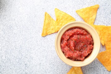 Spicy salsa sauce in bowl and nachos on light grey table, flat lay. Space for text