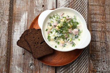 Delicious okroshka soup in bowl and bread on wooden table, top view