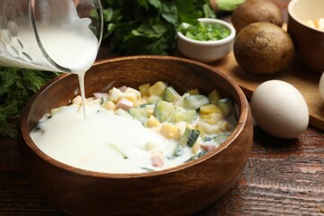 Making okroshka soup. Adding kefir to bowl with ingredients at wooden table, closeup