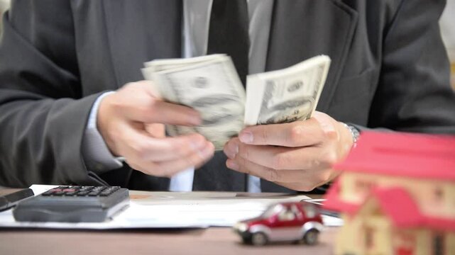 Businessman counting dollar bills at a desk with a calculator, documents, and small toy models of a house and car, symbolizing financial planning, budgeting, or property investment