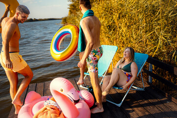 Group of friends in swimsuit sitting on a pier talking and drinking beer enjoying a summer day at the lake.