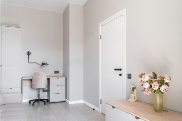 cozy office in light, soft colors. Pleasant lighting, beige walls, desk, comfortable chair, desk lamp. vase of pink flowers on dresser and gray sofa. Floral home decoration. Architecture inside home