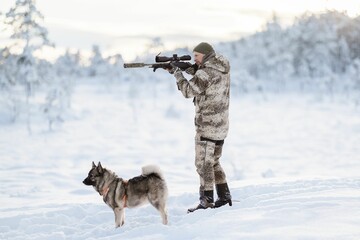 Hunter in camouflage with a rifle and a dog in a snowy landscape