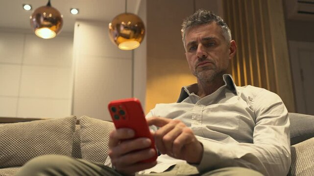 Gray-haired serious elder man is sitting on the couch in business outfit holding his mobile phone in hands and scrolling. He seems focused and thoughtful