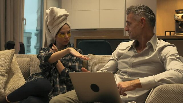 European couple is sitting on a couch in the living room in casual outfit. A beautiful woman with a towel on her head showing her manicure to her boyfriend who is focused on laptop