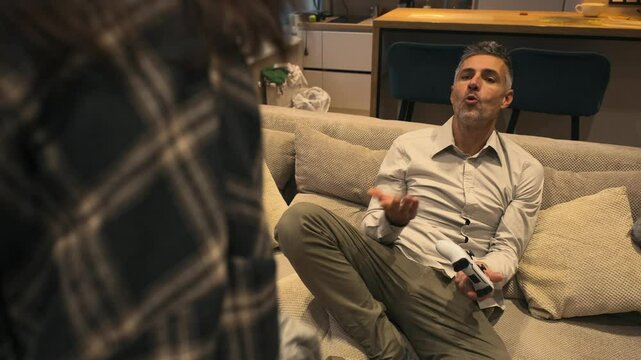 Couple argues. A gray-haired man sits on the couch with a joystick, while his girlfriend throws dirty clothes at him, frustrated that he hasn't cleaned up. She is angrily scolding him