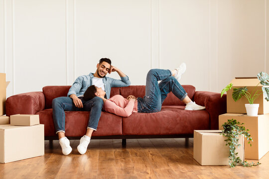Moving Day. Lovely couple relaxing on the couch in new home with cardboard boxes around. Happy man and woman lying on sofa and dreaming, discussing future interior design, free copy space
