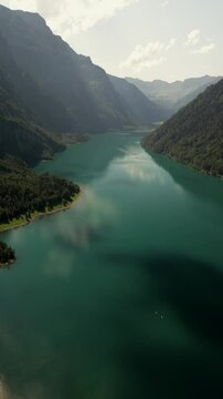 Vertical drone shot of beautiful glacial emerald-green lake surrounded by lush forests, natural lake in summer afternoon under clear blue sky