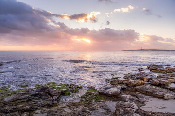 Obraz premium Serene Beauty Of A Rocky Shoreline At Sunset, With Sea Waves Gently Crashing Against The Rocks And A Lighthouse In The Background. The Sky Is Illuminated With Soft Pink And Orange Punta Prima, Menorca