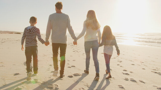 Rear View Of Family Walking On Winter Beach Holding Hands With Flaring Sun