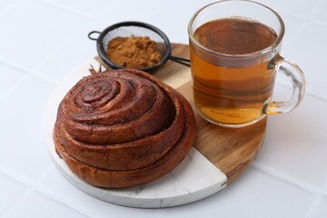 Delicious cinnamon roll bun, tea and spices on white tiled table, closeup