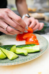 A close-up of hands preparing fresh vegetables, including sliced tomatoes and cucumbers, on a decorative plate. The scene showcases healthy food preparation in a modern kitchen.