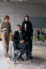 Fototapeta premium A diverse business team poses confidently, including a smiling director seated in a wheelchair, flanked by two professional female colleagues, one wearing a hijab and the other holding a tablet, in a
