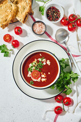 Spanish Cold Tomato Gazpacho Soup garnished with parsley, feta cheese and spices with toasted bread slices, tomatoes and herbs on white table