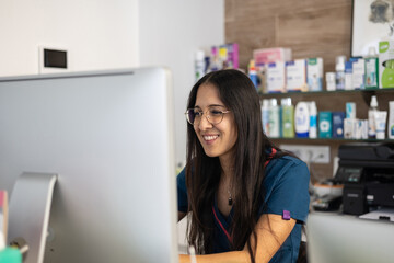 Young veterinarian smiling and working on desktop computer in a veterinary clinic with shelves full of products in the background