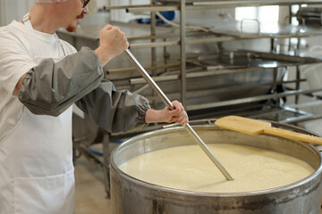 Person stirring large vat of milk in industrial-scale cheese production facility, wearing protective clothing and safety gear surrounded by metal equipment and shelving