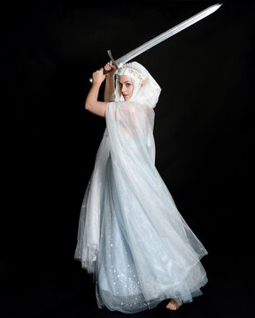 full length portrait beautiful blonde fantasy maiden faery wearing medieval fairytale ballgown costume, flowing white hooded cape. standing pose holding a sword weapon, isolated dark studio background