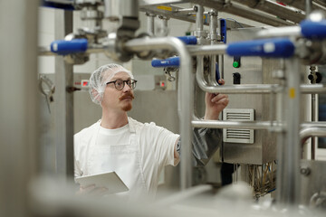 Man examining industrial machinery in food processing plant wearing hairnet and gloves, carefully checking equipment. Holding tablet for data input