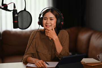 A female podcaster wearing headphones, sitting at a desk with a microphone, tablet, and notes, pausing to think deeply while smiling during her recording session