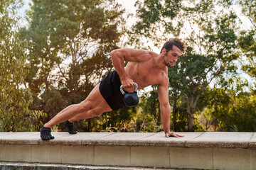 Muscular athlete doing renegade rows with kettlebell in a park