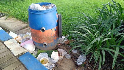 A blue trash can is overflowing with various types of garbage, including plastic bags, food wrappers, and styrofoam containers. The trash is scattered around the can and on the ground, creating a mess