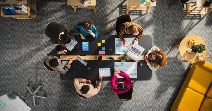Top Down View: Startup Meeting Room with Team of Diverse Entrepreneurs Sitting at the Conference Table to Discuss Problems, Use Tablet and Laptop Computers, Share Documents with Business Statistics - Powered by Adobe