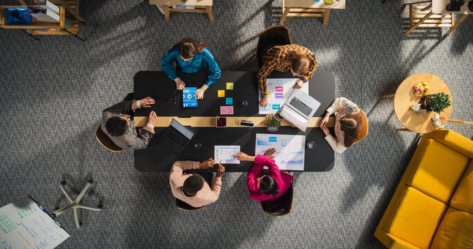 Startup Meeting Room: Team of Entrepreneurs Sitting Around Conference Table, Having Discussions, Solving Business Problems, Using Tablet, Laptop, Share Documents with Statistics, Charts. Top Down View