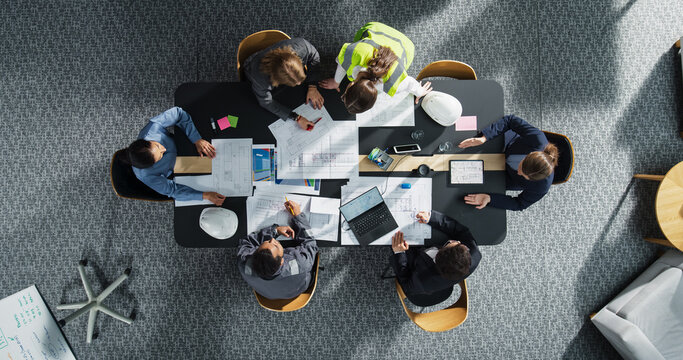Top Down View of Diverse Team of Real Estate Construction Engineers Discussing Project Blueprints in an Office. Architectural Bureau With Colleagues Using Tablet, Laptop, Wearing Reflective Jacket.