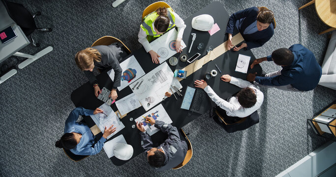 Top Down View of Diverse Team of Real Estate Construction Engineers Discussing Blueprints, Documents in Corporate Office. Modern Workspace of Architectural Bureau With Colleagues Using Tablet, Laptop.